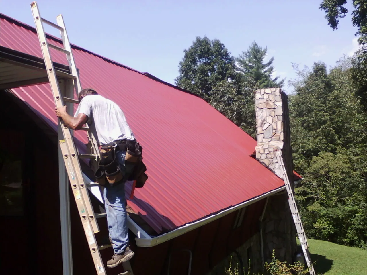 Roofer on a ladder beside a red metal roof and stone chimney — Soffit & Fascia Repair by Roofing Brothers in Mokelumne Hill